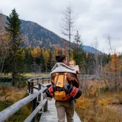 Ojciec z dzieckiem na plecach spaceruje drewnianą kładką pośród jesiennych lasów. Tatry z dzieckiem to piękna przygoda.