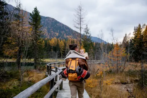 Ojciec z dzieckiem na plecach spaceruje drewnianą kładką pośród jesiennych lasów. Tatry z dzieckiem to piękna przygoda.